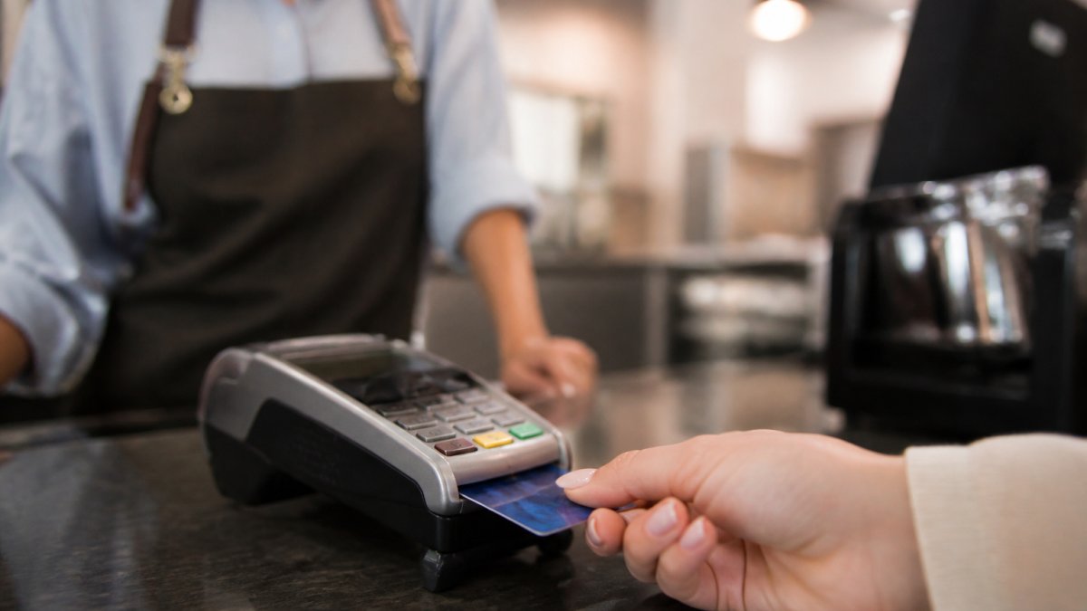 Close-up of a customer's hand inserting a credit card into a point-of-sale terminal at a retail counter, with the merchant and terminal visible in the background